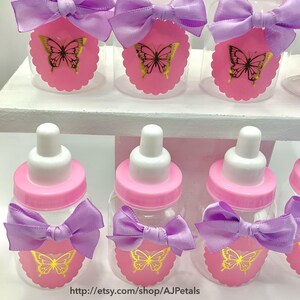 Puede incluir: Una colecci&oacute;n de biberones de pl&aacute;stico transparente con tapas de rosca rosas y tetinas blancas. Cada biber&oacute;n presenta un lazo de sat&eacute;n lavanda y una etiqueta de papel rosa festoneada decorada con un gr&aacute;fico de mariposa dorada. El texto http://etsy.com/shop/AJPetals es visible en la parte inferior.