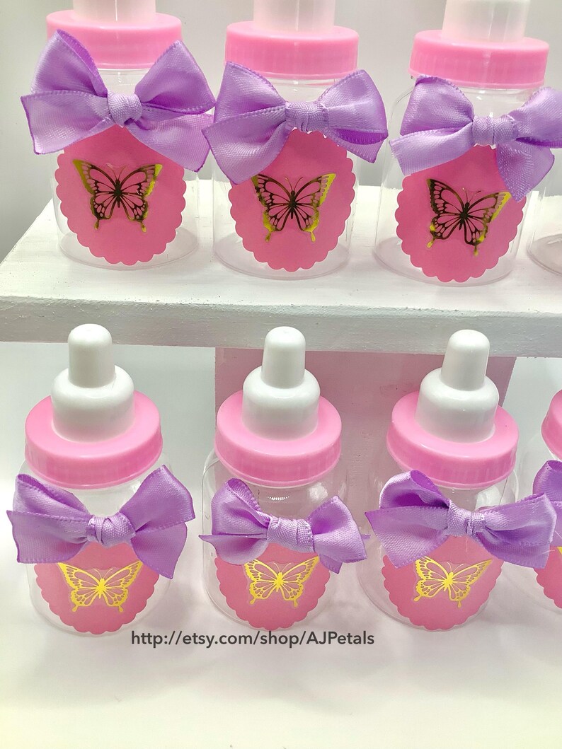 Puede incluir: Una colecci&oacute;n de biberones de pl&aacute;stico transparente con tapas de rosca rosas y tetinas blancas. Cada biber&oacute;n presenta un lazo de sat&eacute;n lavanda y una etiqueta de papel rosa festoneada decorada con un gr&aacute;fico de mariposa dorada. El texto http://etsy.com/shop/AJPetals es visible en la parte inferior.