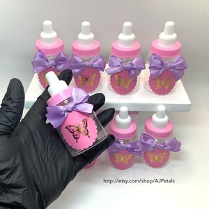 Puede incluir: Un conjunto de biberones de pl&aacute;stico transparente con tapas rosas y tetinas blancas. Cada biber&oacute;n tiene una etiqueta rosa festoneada con un gr&aacute;fico de mariposa dorada y un lazo de sat&eacute;n color lavanda atado alrededor del cuello del biber&oacute;n.