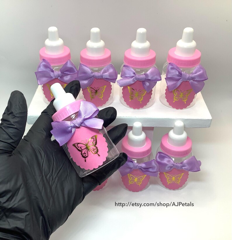 Puede incluir: Un conjunto de biberones de pl&aacute;stico transparente con tapas rosas y tetinas blancas. Cada biber&oacute;n tiene una etiqueta rosa festoneada con un gr&aacute;fico de mariposa dorada y un lazo de sat&eacute;n color lavanda atado alrededor del cuello del biber&oacute;n.
