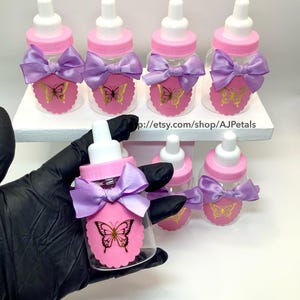 Puede incluir: Biberones de pl&aacute;stico transparente con tetinas blancas y tapas rosas de rosca. Cada biber&oacute;n presenta un lazo de sat&eacute;n lavanda y una etiqueta rosa festoneada con un gr&aacute;fico de mariposa dorada. El texto p://etsy.com/shop/AJPetals es visible.