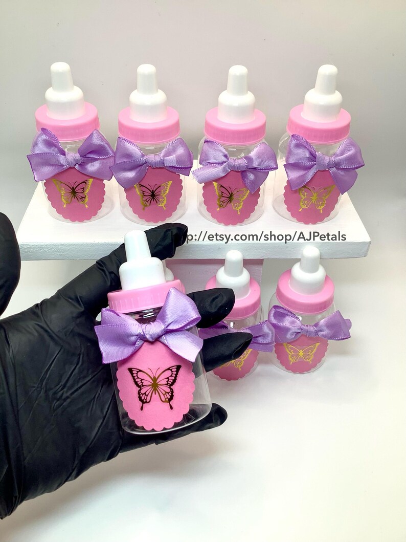 Puede incluir: Biberones de pl&aacute;stico transparente con tetinas blancas y tapas rosas de rosca. Cada biber&oacute;n presenta un lazo de sat&eacute;n lavanda y una etiqueta rosa festoneada con un gr&aacute;fico de mariposa dorada. El texto p://etsy.com/shop/AJPetals es visible.