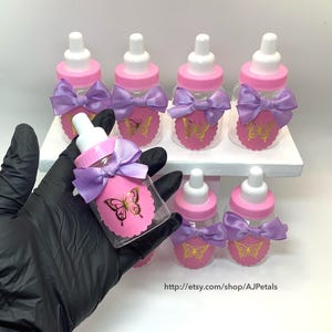 Puede incluir: Un conjunto de biberones de pl&aacute;stico transparente con tapas rosas y tetinas blancas. Cada biber&oacute;n tiene una etiqueta rosa festoneada con un gr&aacute;fico de mariposa dorada y un lazo de sat&eacute;n color lavanda atado alrededor del cuello del biber&oacute;n.