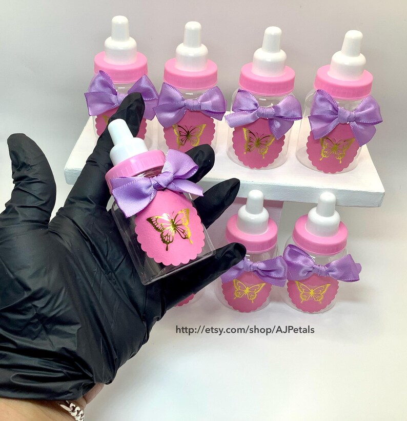 Puede incluir: Un conjunto de biberones de pl&aacute;stico transparente con tapas rosas y tetinas blancas. Cada biber&oacute;n tiene una etiqueta rosa festoneada con un gr&aacute;fico de mariposa dorada y un lazo de sat&eacute;n lavanda. El texto dice http://etsy.com/shop/AJPetals.