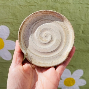 May include: A handmade ceramic spoon rest with a spiral design. The spoon rest is beige with a brown rim and a small lip for resting utensils. The background is a green wall with white daisy cutouts.