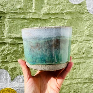 May include: A cylindrical ceramic planter with a speckled beige base and a smooth, glossy glaze that transitions from a deep teal and forest green at the bottom to a lighter, muted white and grey tone at the top rim, featuring a wide opening.