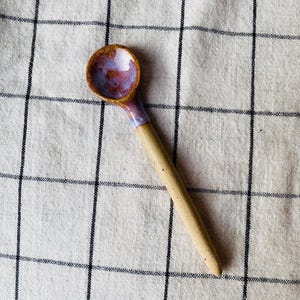 May include: A handcrafted ceramic spoon with a circular bowl and a long handle. The bowl features a purple and brown glaze, and the handle is a speckled beige. The spoon rests on a white and black checked cloth.