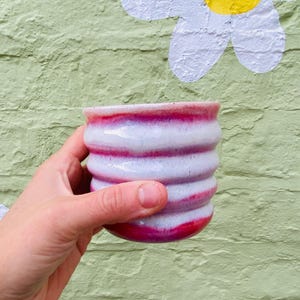 May include: A ceramic cup with a ribbed, cylindrical body featuring a horizontal striped pattern in alternating bands of glossy magenta pink and opaque white glaze, with a smooth, rounded rim and a slightly tapered base design.
