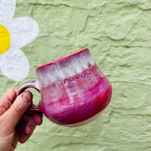 May include: A ceramic mug with a rounded body and a curved handle. The glaze features a gradient transition from a deep magenta at the base to a lighter pink and white at the rim, accented by a horizontal band of small, raised reddish-brown dots.