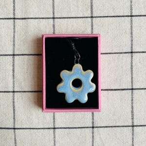 May include: A pale blue ceramic flower pendant, presented in a pink box with a black interior. The pendant is suspended from a black cord. The background is a checkered fabric.