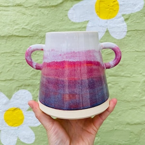 May include: A handcrafted ceramic vase with two loop handles. The vase displays a colour gradient, beginning with white at the top, shifting through pink and red, and finishing with a deep blue at the base. The base is a light beige colour.