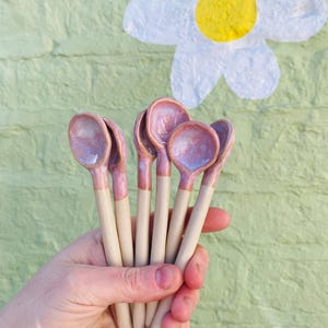 May include: A hand holds six ceramic spoons. The spoon bowls are a glossy pinkish-purple, with a matching band at the base of the handle. The handles are a light beige colour. The background is a light green wall with a white and yellow daisy painted on it.