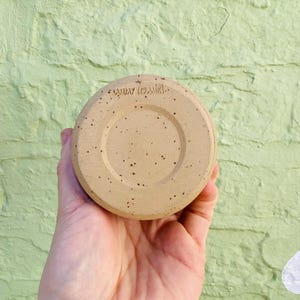 Peut inclure: Une soucoupe en c&eacute;ramique beige avec un motif tachet&eacute; et le texte "SUNDAY CERAMICS" estampill&eacute; au fond. La soucoupe est ronde avec un centre en retrait et est tenue dans la main d'une personne.