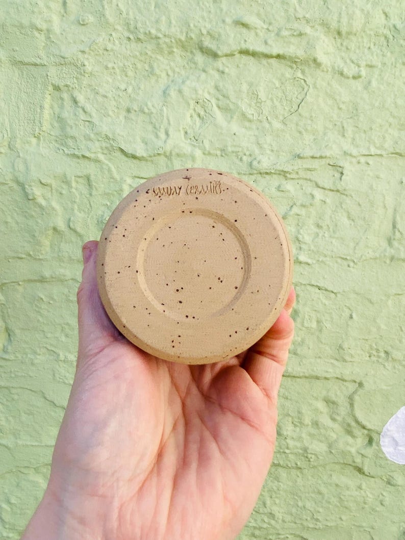 Peut inclure: Une soucoupe en c&eacute;ramique beige avec un motif tachet&eacute; et le texte "SUNDAY CERAMICS" estampill&eacute; au fond. La soucoupe est ronde avec un centre en retrait et est tenue dans la main d'une personne.