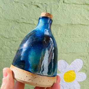 Peut inclure: Une bouteille en c&eacute;ramique faite &agrave; la main avec un bouchon en li&egrave;ge. La bouteille pr&eacute;sente un d&eacute;grad&eacute; de gla&ccedil;ure bleu fonc&eacute; et sarcelle avec des accents mouchet&eacute;s. La base est non &eacute;maill&eacute;e, r&eacute;v&eacute;lant une couleur d'argile naturelle et mouchet&eacute;e.