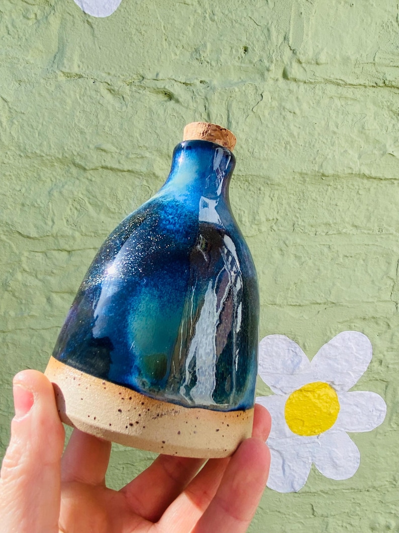 Peut inclure: Une bouteille en c&eacute;ramique faite &agrave; la main avec un bouchon en li&egrave;ge. La bouteille pr&eacute;sente un d&eacute;grad&eacute; de gla&ccedil;ure bleu fonc&eacute; et sarcelle avec des accents mouchet&eacute;s. La base est non &eacute;maill&eacute;e, r&eacute;v&eacute;lant une couleur d'argile naturelle et mouchet&eacute;e.