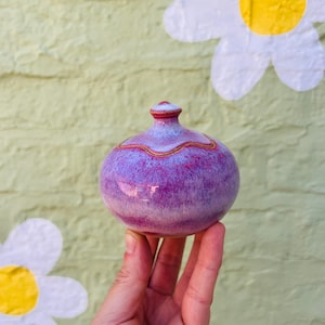 May include: A small, round ceramic jar with a lid, featuring a vibrant purple and magenta glaze with a smooth, glossy finish. The jar has a narrow neck and a small, rounded knob on top of the lid, with a visible seam where the lid meets the base.