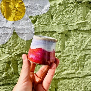 May include: A small ceramic cup featuring a vibrant gradient glaze that transitions from a deep, saturated magenta at the base to a soft, speckled white and light blue at the rim. The cup has a smooth, glossy finish and a slightly tapered shape.