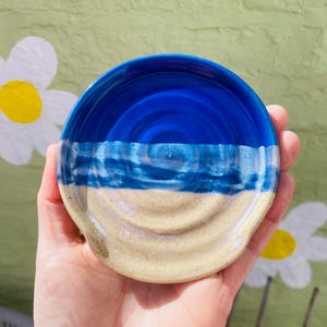 May include: A ceramic spoon rest, featuring a blue and beige design. The top half is a deep blue, fading to a lighter blue and then beige. The spoon rest has a curved form with a small lip, ideal for resting kitchen utensils.
