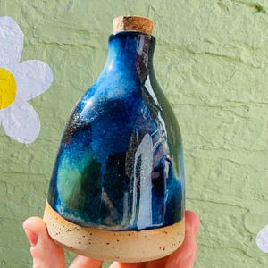 Peut inclure: Une bouteille en c&eacute;ramique faite &agrave; la main avec un bouchon en li&egrave;ge. La bouteille pr&eacute;sente un gla&ccedil;age bleu fonc&eacute; et vert et une base tachet&eacute;e de couleur beige. La bouteille est tenue dans une main sur un fond vert clair textur&eacute; avec des d&eacute;coupes de fleurs blanches.