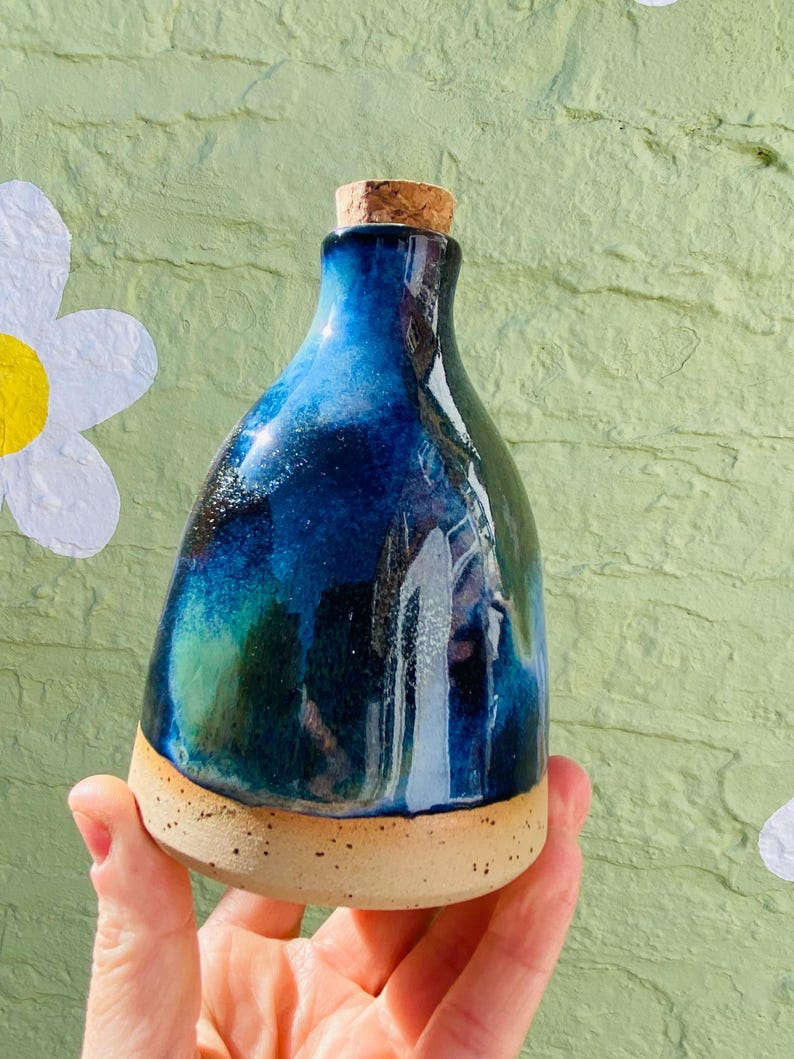 Peut inclure: Une bouteille en c&eacute;ramique faite &agrave; la main avec un bouchon en li&egrave;ge. La bouteille pr&eacute;sente un gla&ccedil;age bleu fonc&eacute; et vert et une base tachet&eacute;e de couleur beige. La bouteille est tenue dans une main sur un fond vert clair textur&eacute; avec des d&eacute;coupes de fleurs blanches.
