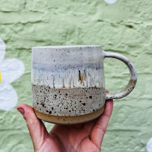 May include: A ceramic mug featuring a speckled finish with a white glazed upper section that transitions into a textured, tan-colored base. The glaze displays vertical drip patterns where the white meets the speckled clay, and a curved handle.