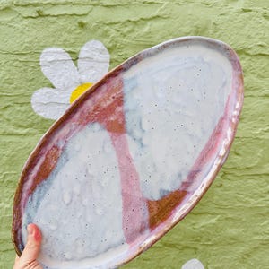 May include: An oval ceramic platter with an irregular, hand-formed edge. The surface features a speckled white glaze accented by two distinct, vertical bands of muted pink and earthy brown tones that create a segmented, abstract colour pattern.