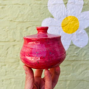 May include: A small, round, handmade ceramic jar with a matching lid, featuring a vibrant, glossy magenta-pink glaze with subtle dark speckles throughout the surface. The container has a smooth, curved silhouette and a small knob on the lid.