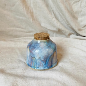 May include: A small ceramic jar with a cork stopper. The jar is glazed in shades of blue, purple, and white. The cork stopper is light brown. The jar has a rounded shape. The jar is sitting on a white surface.