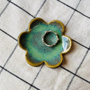 May include: A handmade ceramic flower-shaped ring dish with a turquoise and brown glaze. A silver ring rests in the centre of the dish. The dish is on a white and black checkered cloth.