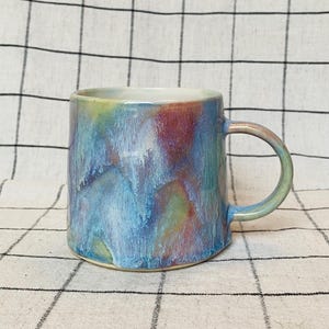 May include: A ceramic mug with a colourful, abstract glaze in shades of blue, purple, and green. The mug has a rounded handle and a square base. The interior is white.