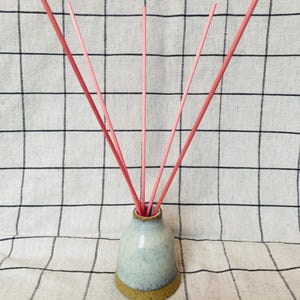 May include: A small, light blue ceramic essential oil diffuser with a tan base. Pink reeds are inserted into the diffuser. The diffuser sits on a white and black checked cloth.