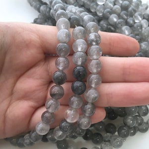 Könnte beinhalten: Stränge grauer Edelsteinperlen. Die Perlen sind rund und haben eine durchscheinende Qualität, mit Farbvariationen von hellgrau bis dunkelgrau. Die Perlen werden in einer Hand gehalten, wobei weitere Stränge im Hintergrund sichtbar sind.