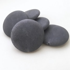 May include: Five smooth, round, gray stones with a hole drilled through each. The stones are arranged in a pile.