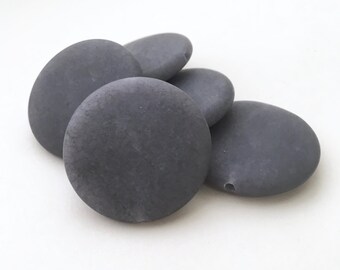 May include: Five smooth, round, gray stones with a hole drilled through each. The stones are arranged in a pile.
