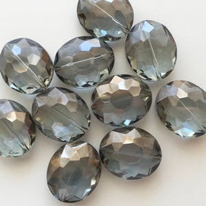 May include: A group of ten oval-shaped faceted glass beads in a dark gray color. The beads are scattered on a white surface.