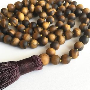 May include: A brown and gold tiger's eye mala bead necklace with a dark purple tassel. The necklace is made of round beads and is approximately 108 beads long.