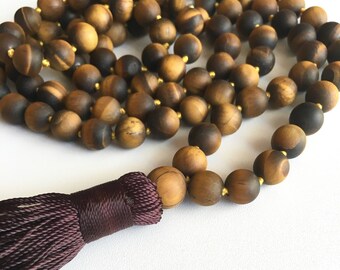 May include: A brown and gold tiger's eye mala bead necklace with a dark purple tassel. The necklace is made of round beads and is approximately 108 beads long.