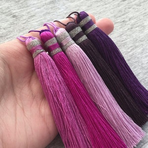 Könnte beinhalten: Fünf bunte Quasten in Rosatönen, Lila und Lavendel. Die Quasten bestehen aus weichen, seidenartigen Fäden und haben einen silbernen Besatz oben.