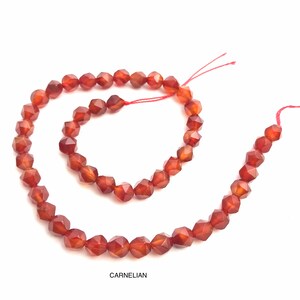 May include: Strands of faceted carnelian beads in shades of orange and brown. The beads are strung on a red thread, with the word "CARNELIAN" printed below.
