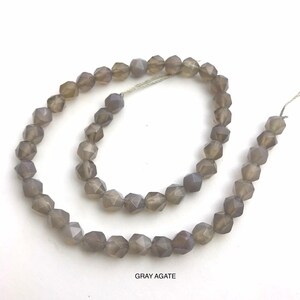 May include: A strand of faceted gray agate beads. The beads are a muted gray color and have a geometric, multi-faceted shape. The beads are strung on a thin, silver-colored thread. The text "GRAY AGATE" is at the bottom of the image.