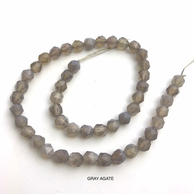 May include: A strand of faceted gray agate beads. The beads are a muted gray color and have a geometric, multi-faceted shape. The beads are strung on a thin, silver-colored thread. The text "GRAY AGATE" is at the bottom of the image.