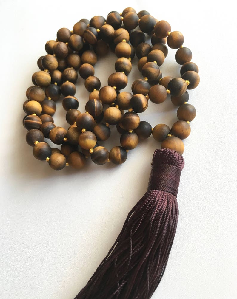 May include: A brown tiger's eye gemstone mala with a dark brown tassel. The mala is made of round beads and is strung on a gold cord.