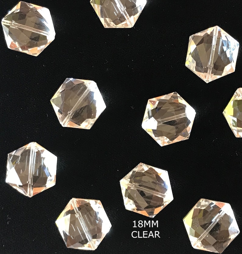 May include: A close-up of ten clear glass faceted hexagon beads, each measuring 18mm in diameter. The beads are arranged on a black surface. 18MM CLEAR