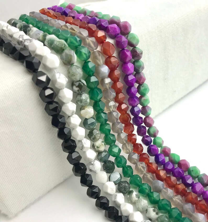 May include: A collection of faceted gemstone beads in various colors, including black, white, green, red, purple, and gray. The beads are strung together, showcasing their geometric shapes and natural variations. Ideal for jewelry making.