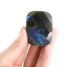 May include: A faceted, rectangular labradorite gemstone with iridescent blue, green, and gold flashes. The stone is held in a hand against a white background, showcasing its polished surface and unique color play. This gemstone is ideal for jewelry making.