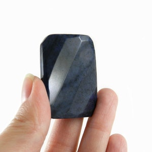 May include: A dark blue, faceted, gemstone with a smooth, polished surface. The gemstone is held between two fingers.