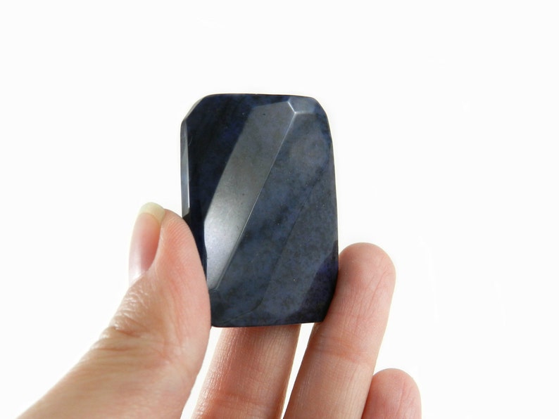 May include: A dark blue, faceted, gemstone with a smooth, polished surface. The gemstone is held between two fingers.