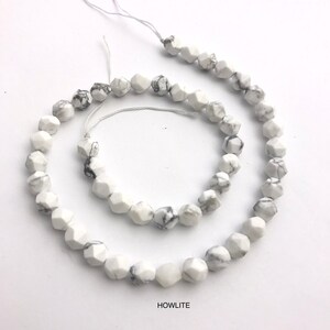 May include: Strand of white howlite gemstone beads with gray veining. The beads are faceted, displaying a geometric shape. The word "HOWLITE" is printed in black at the bottom of the image. The beads are strung on a white string.