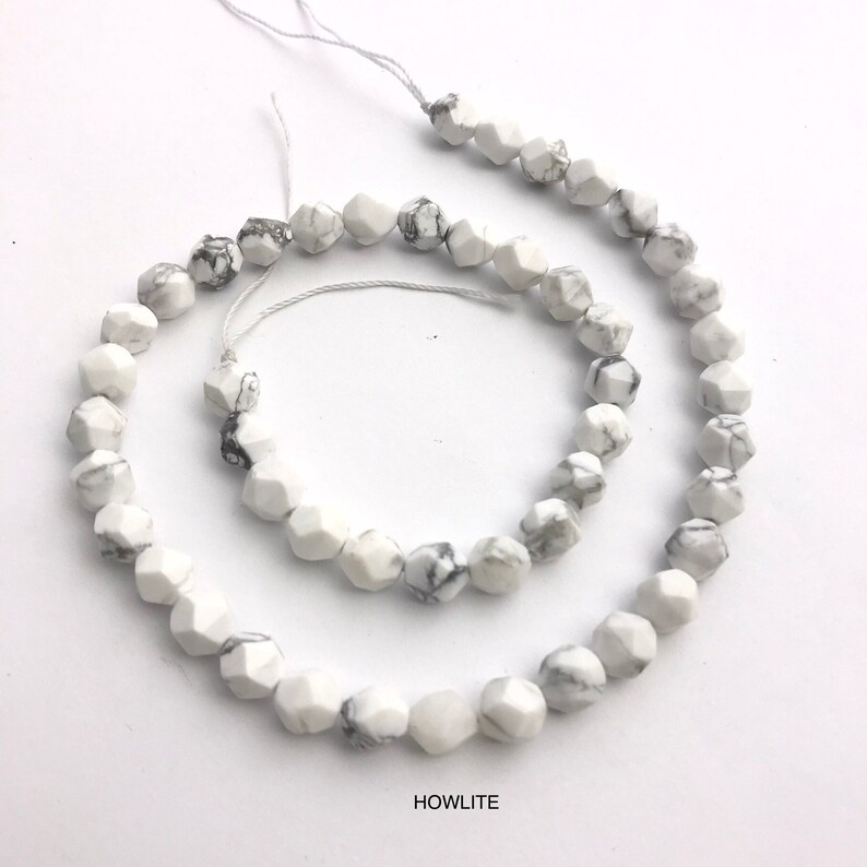 May include: Strand of white howlite gemstone beads with gray veining. The beads are faceted, displaying a geometric shape. The word "HOWLITE" is printed in black at the bottom of the image. The beads are strung on a white string.
